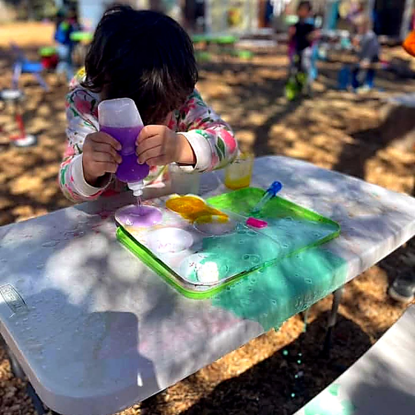 A child mixing coloured liquids in a tray at an outdoor table