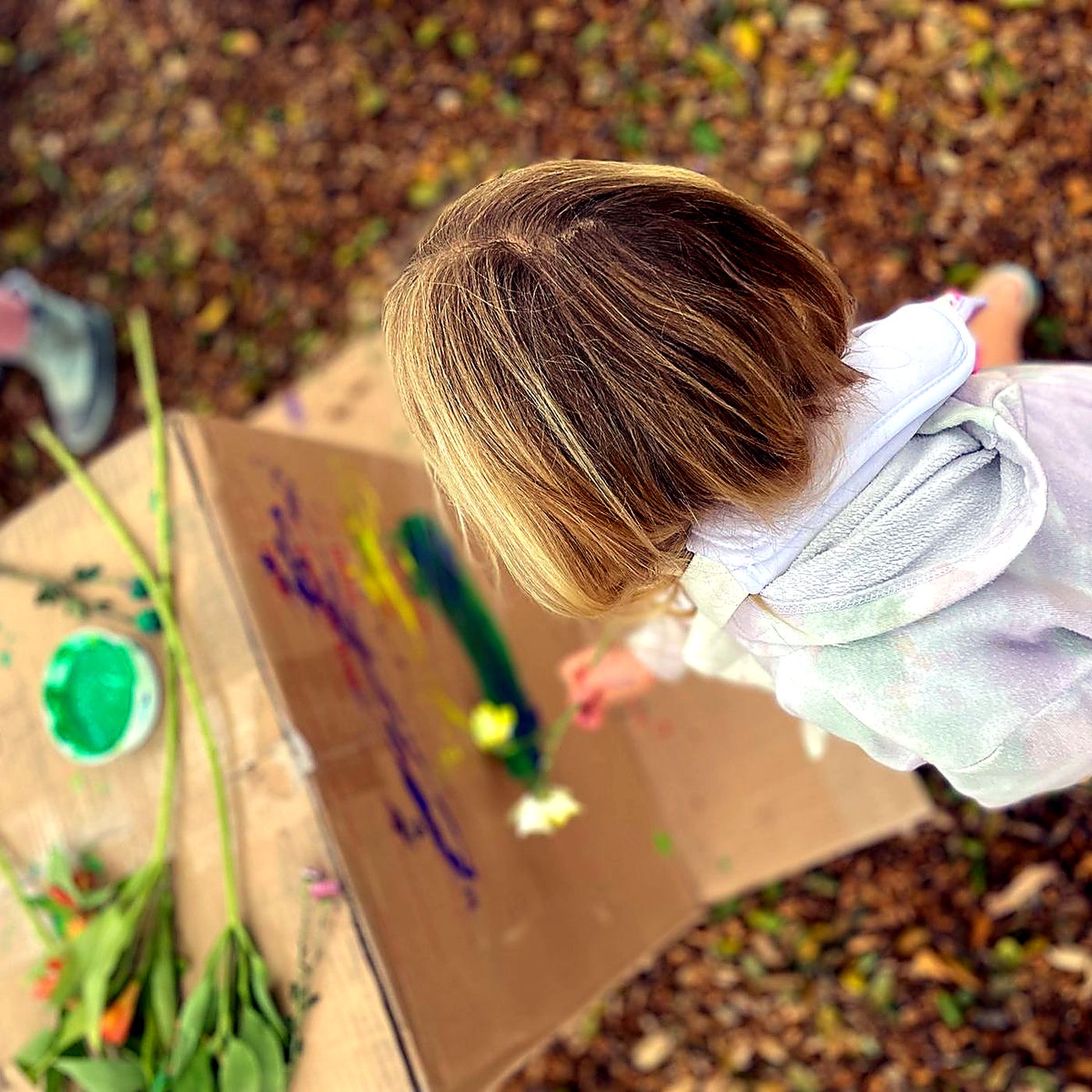 A child painting on cardboard outdoors, surrounded by flowers and natural materials