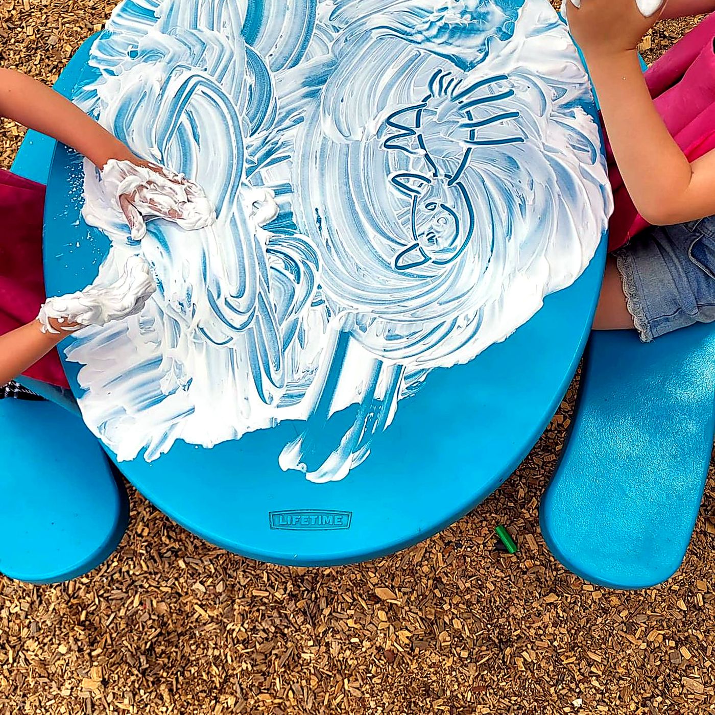 Children making swirling patterns in shaving foam on an outdoor table